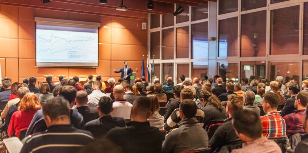 A large group of people sitting down, listening to a presentation. The presenter points at a graph on the projector.